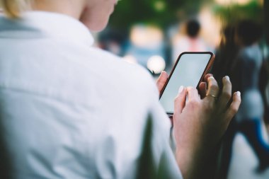 Arka planda, kısa saçlı, beyaz gömlekli, parkta vakit geçirirken modern cep telefonunun ekranına dokunan tanınmamış bir kadın var.