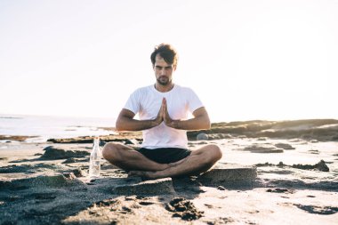 Spor kıyafetli güçlü bir erkek Lotus pozunda yoga yapıyor ve göğsüne yakın bir yerde Namaskara Mudra hareketi yapıyor.