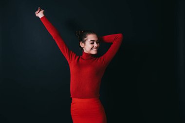 Kırmızı elbiseli neşeli, zayıf esmer gözleri kapalı, elleri arkada stüdyoda koyu gri arka planda duruyor.