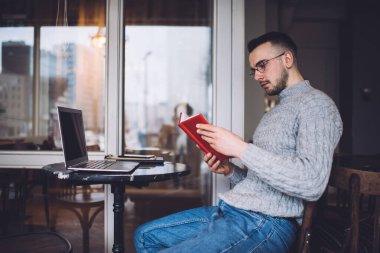 Serious young male worker wearing warm knitted sweater and eyeglasses sitting at table with book in hands while working remotely from home