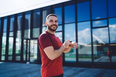 Mutlu erkek, güneşli havada modern cam binanın bulanık arka planında dururken kameraya bakıp spor bilekliğine dokunuyor.