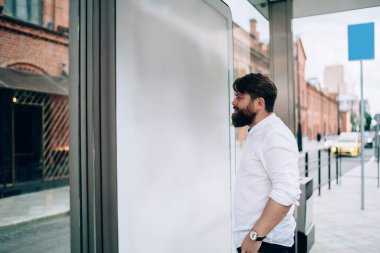 Gündüz vakti modern şehirde otobüs durağında beyaz tişörtlü genç sakallı bir adamın yan görüntüsü.