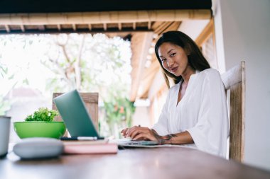 Zarif beyaz elbiseli çekici bir Asyalı kadın ahşap masada oturuyor ve gündüz vakti online çalışırken dizüstü bilgisayarını inceliyor.