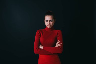 Ciddi bir kadın model, elleri katlı kameraya bakarken somurtuyor ve somurtuyor stüdyoda siyah duvarın yanında duruyor.