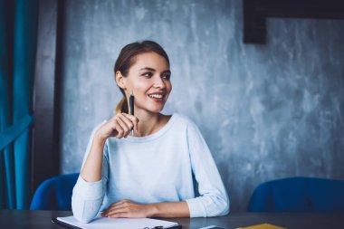 Modern işyerindeki ahşap masada oturan ve başka tarafa bakan, günlük kıyafetli, neşeli, genç bir kadın serbest yazar.