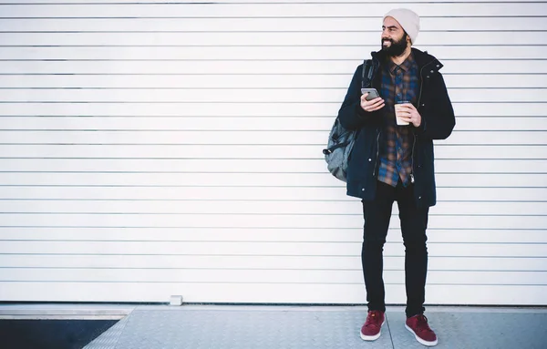 Casual dressed hipster backpacker with modern cellphone technology and caffeine beverage in hands looking away