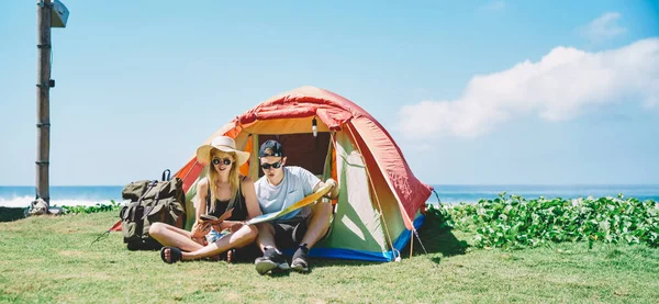 Full body couple travelers checking map while sitting by camping tent with sea in background in sunny summer day with blue sky
