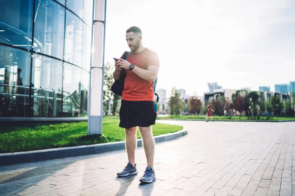 Full body of bearded male in sport clothing messaging on mobile phone while standing near modern building in city on pavement in sunlight