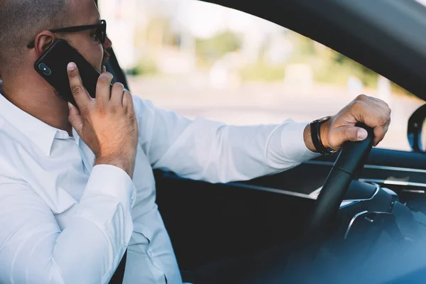 Side view of male executive in white shirt sitting in modern vehicle while discussing business project on smartphone during work