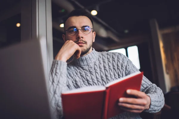 在现代工作场所 穿着保暖毛衣和眼镜的大胡子男性一边在笔记本电脑上工作 一边看着相机 一边看书 — 图库照片
