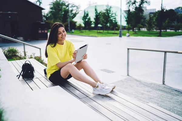 Full body of happy ethnic female in casual clothes sitting on wooden podium in park and smiling while browsing tablet