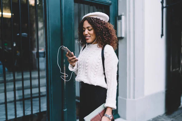 暗い肌のトレンディーな服装をした女性が携帯電話を持って街を歩く笑顔プレイリスト音楽を選択 — ストック写真