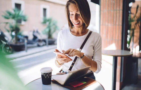 路上の小さなテーブルでコーヒーと一緒に立っている間幸せな笑顔で携帯電話を使用してカジュアルな服装で中年女性 — ストック写真