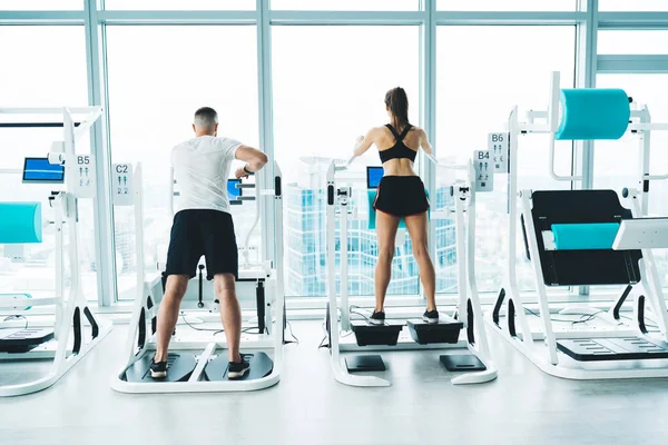 Back view full length of anonymous slim sportspeople in activewear training on simulators with digital screen during workout in spacious gym