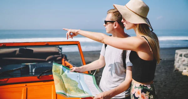 Side view of young couple standing near car with map looking and pointing away choosing way while travelling together during summer holidays