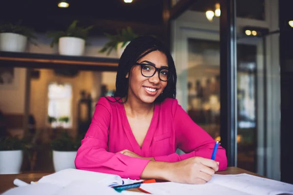 Happy black female journalist with braids in casual wear and glasses looking at camera ad smiling while working on article in creative workspace