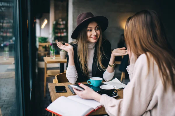Şapkalı olumlu bir kadın, bayan arkadaşıyla aynı masada oturuyor ve modern restoranda konuşurken el kol hareketi yapıyor.