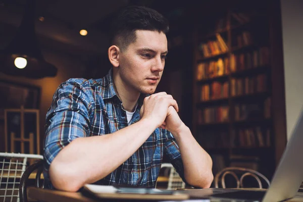 Mavi gömlekli, ciddi bir erkek, elleri sımsıkı bağlı bir şekilde masada oturuyor ve proje üzerinde çalışırken dizüstü bilgisayarında belgeler okuyor.