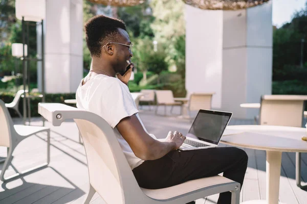 Side view of African American man in casual wear and glasses sitting with laptop on knees while browsing smartphone on terrace in sunny day