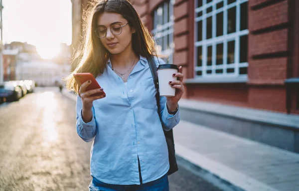 Gözlüklü, vizyon koruması olan beyaz bir kadın cep telefonundan sokaklarda boş zaman geçirerek mesaj okuyor.