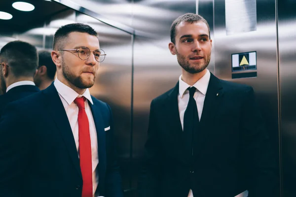 現代のオフィスエレベーターに立つネクタイと眼鏡を着用し 離れて話をしながら見て若いUnshavenビジネスマン — ストック写真