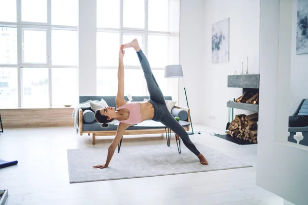 Young strong female with athletic body standing in asana stretching muscles and looking up in apartment,