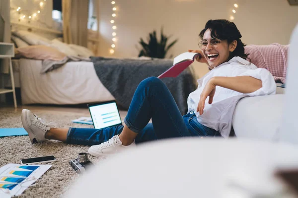 Full body side view of laughing young female freelancer sitting with netbook during remote work in cozy bedroom with glowing garlands