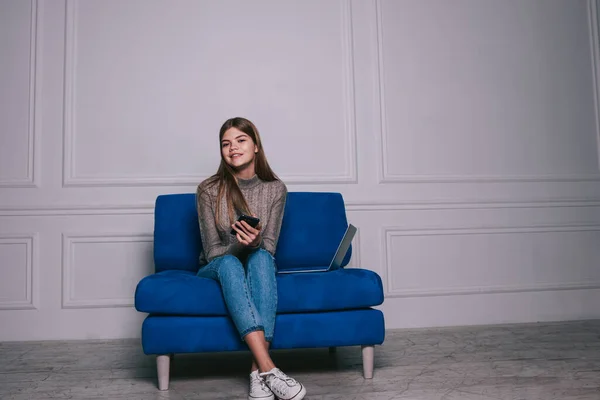 Full body of happy female remote worker in casual outfit with mobile phone and looking at camera while sitting on comfortable sofa