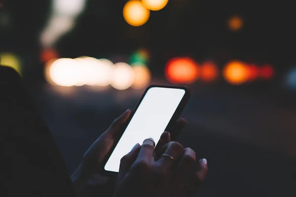 Faceless unrecognizable woman tapping with finger empty blank screen of modern smartphone while standing outdoors with bokeh background at night