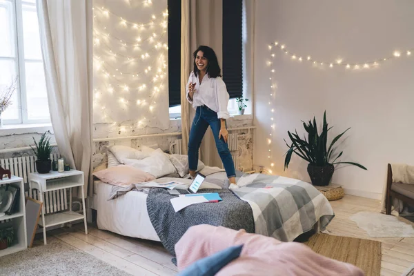 Full body of happy female dancing on bed with smartphone in hand and listening to music in earphones in bedroom with lights on walls