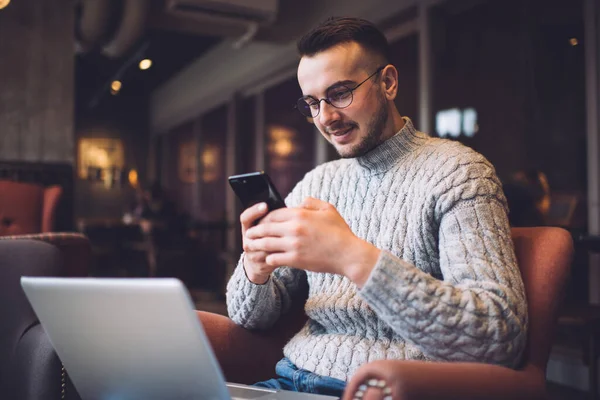 Low angle of content bearded male in casual wear and glasses browsing smartphone while working on laptop in cozy cafe
