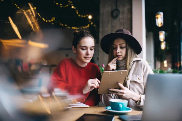 Şapkalı ve günlük kıyafetli genç bir kadın kahve içip kitap okurken gündüzleri kafede oturuyor.