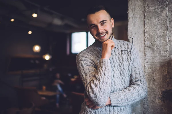 Cheerful bearded male wearing casual warm clothes standing near gray wall holding eyeglasses and looking at camera with toothy smile