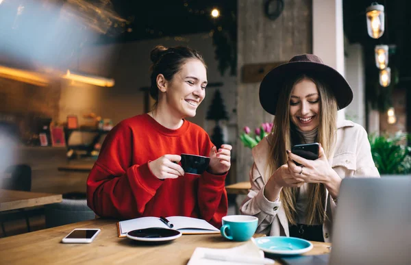 Young female friends in casual outfits sitting at table with cup of coffee and browsing smartphone while spending time together