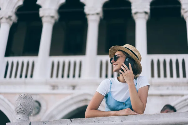 Low angle of trendy female explorer standing near white columns while leaning on railing and listening to partner on cellphone