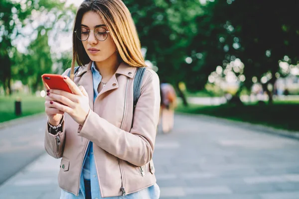 Beautiful millennial woman in eyewear reading messages and notifications received on mobile phone