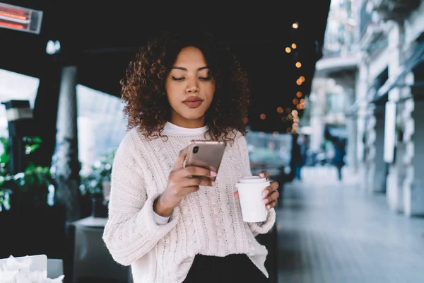若いアフリカ系アメリカ人女性がカジュアルな服装で通りのカフェに立っており 携帯電話を手にテイクアウトコーヒーとブラウジング — ストック写真