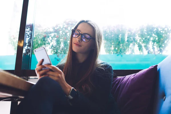 Gözlüklü sakin bir kadın kafeteryadaki yastıklarla kanepede oturuyor ve teneffüste cep telefonu uyarılarını kontrol ederken dinleniyor.