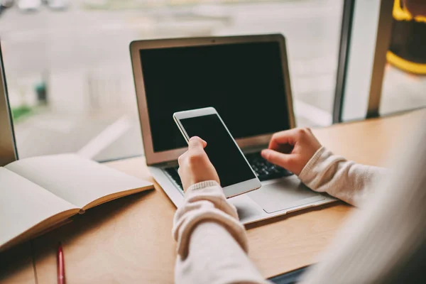 Crop anonymous person in long sleeve sitting at table near window with laptop and notebook browsing smartphone while working in modern workspace