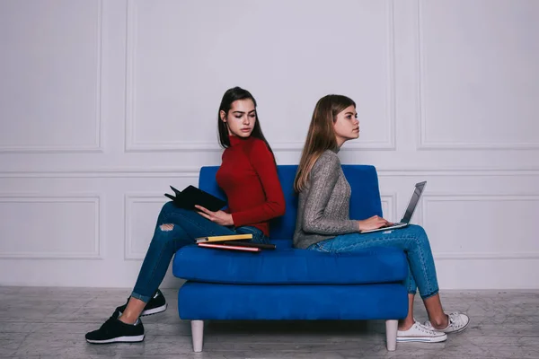 Side view of full body pensive female students in casual outfit looking away while sitting back to back on blue sofa