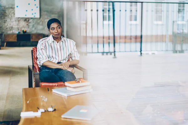 Through glass calm adult African American female in striped formal shirt and denim pants looking away with interest and contemplating while sitting