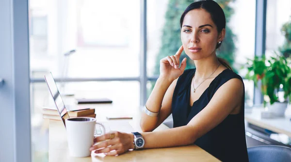 Pensive female entrepreneur with dark hair thinking about business strategy and looking away while sitting at table with laptop in contemporary workplace