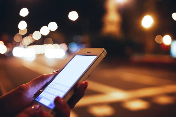 Crop anonymous female using mobile phone while texting message and standing in street on background bokeh light in night city