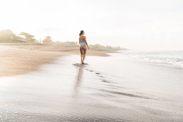 Back view full length graceful slender female in trendy swimwear strolling on wet sandy coast and admiring peaceful sea in tropical resort