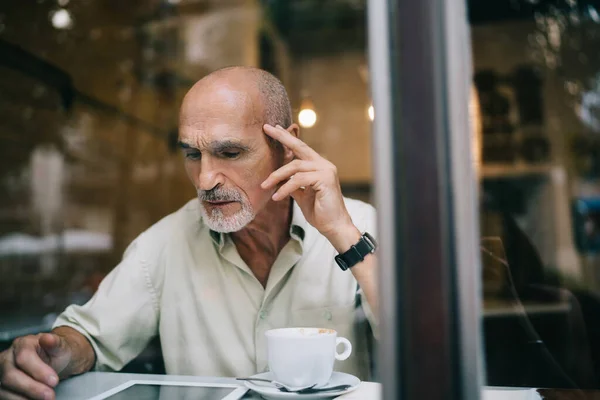 Window view of aged Caucasian male user connecting to public internet in coffee shop for browsing web information via digital tablet gadget