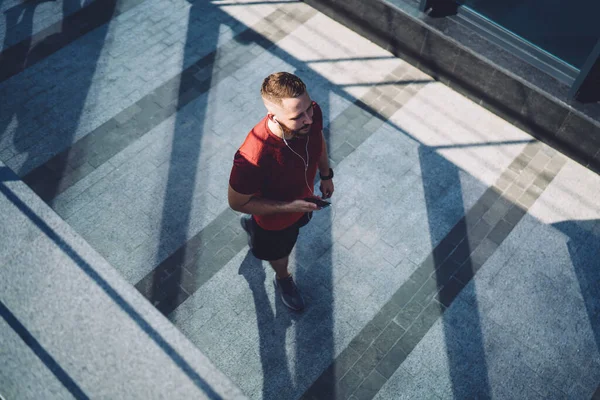 From above of full body male athlete listening to music on earphones and cellphone while looking away and walking after run