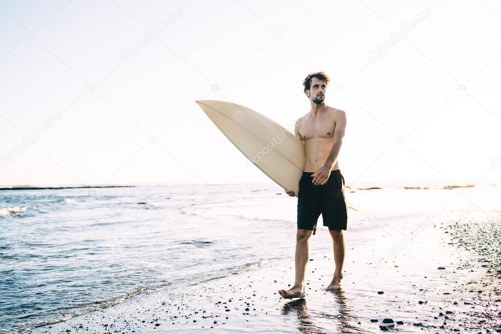 Full body muscular serious male surfer carrying surfboard while strolling along sandy seashore and looking away during sunny clear day