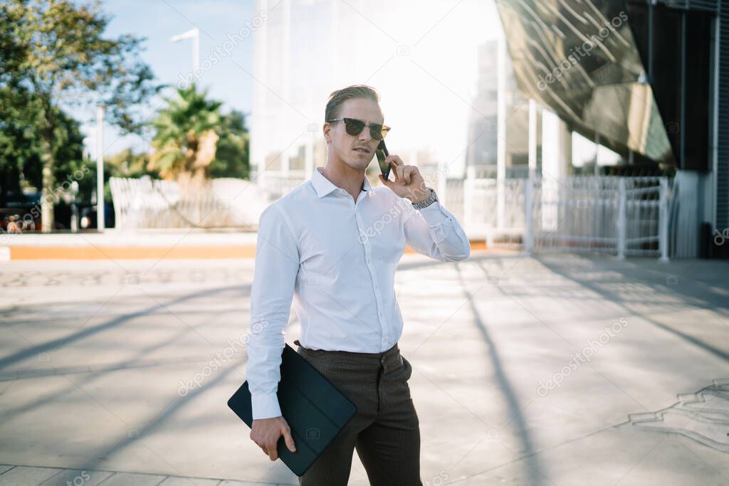 Serious male entrepreneur in formal wear and sunglasses communicating on phone while standing on street near modern building on sunny day