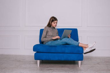 Kazaklı, spor ayakkabılı, netbook 'ta gezinen ve ışık odasındaki rahat koltukta oturan kadın uzaktan kumandalı tam vücut.