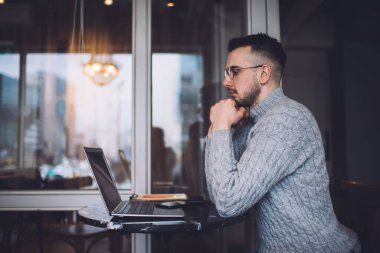 Side view of serious bearded male employee browsing netbook while sitting at table in cozy cafe and working remotely on project